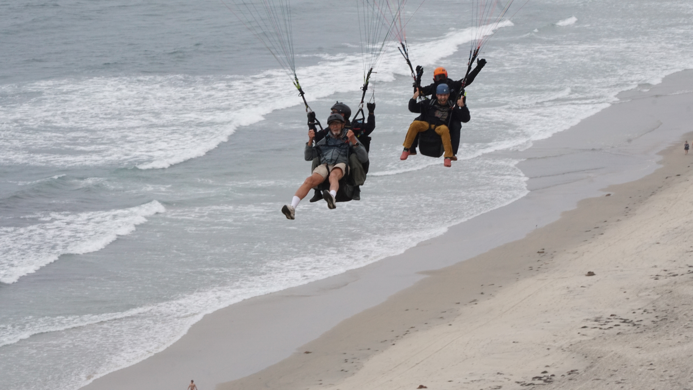 Two people paragliding in tandem along an ocean coastline.
